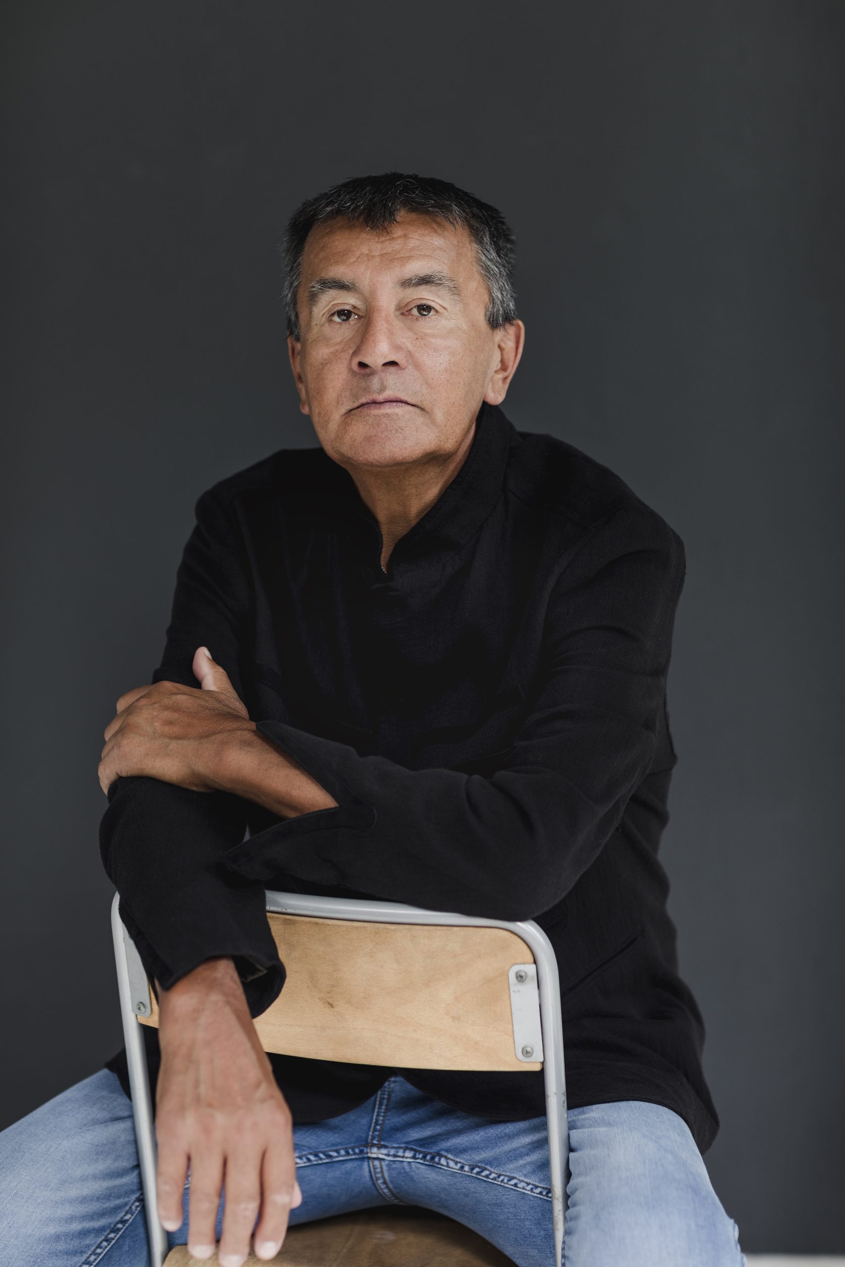 Georges Pisimopeo, un auteur autochtone, est assis sur une chaise, il poste une chemise noire et un jeans. Il regarde l'objectif d'un air sérieux et calme.